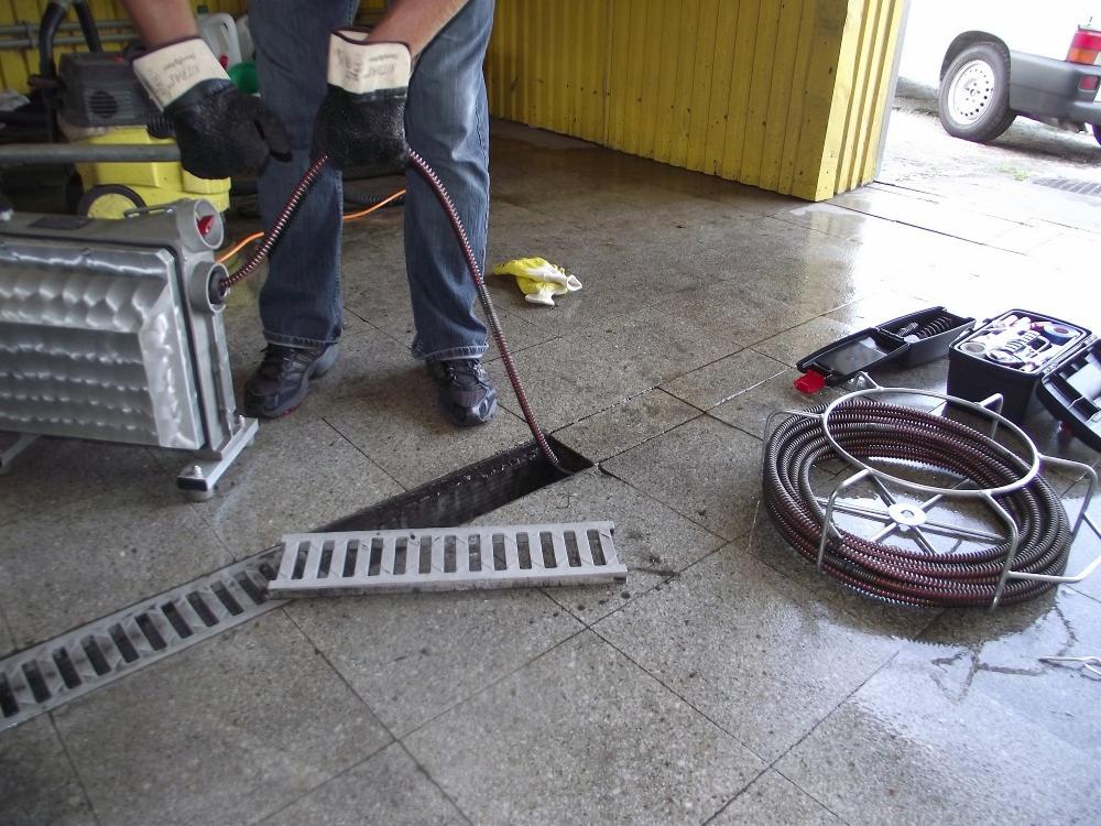 Hydraulik w rękawicach wprowadza sprężynę do czyszczenia kanalizacji podłogowej, obok leży zwinięta sprężyna w stojaku i otwarte skrzynki z narzędziami.