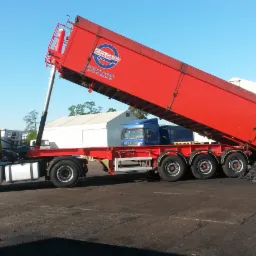 Żółta ciężarówka z czerwoną naczepą wyładowuje węgiel na betonowy plac; widoczny fragment logo producenta naczep.