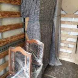 Wnętrze furgonetki z zabezpieczonymi meblami: krzesła owinięte folią stretch, szafa okryta szarym kocem transportowym i spięta sznurkiem, drewniane ściany pojazdu.