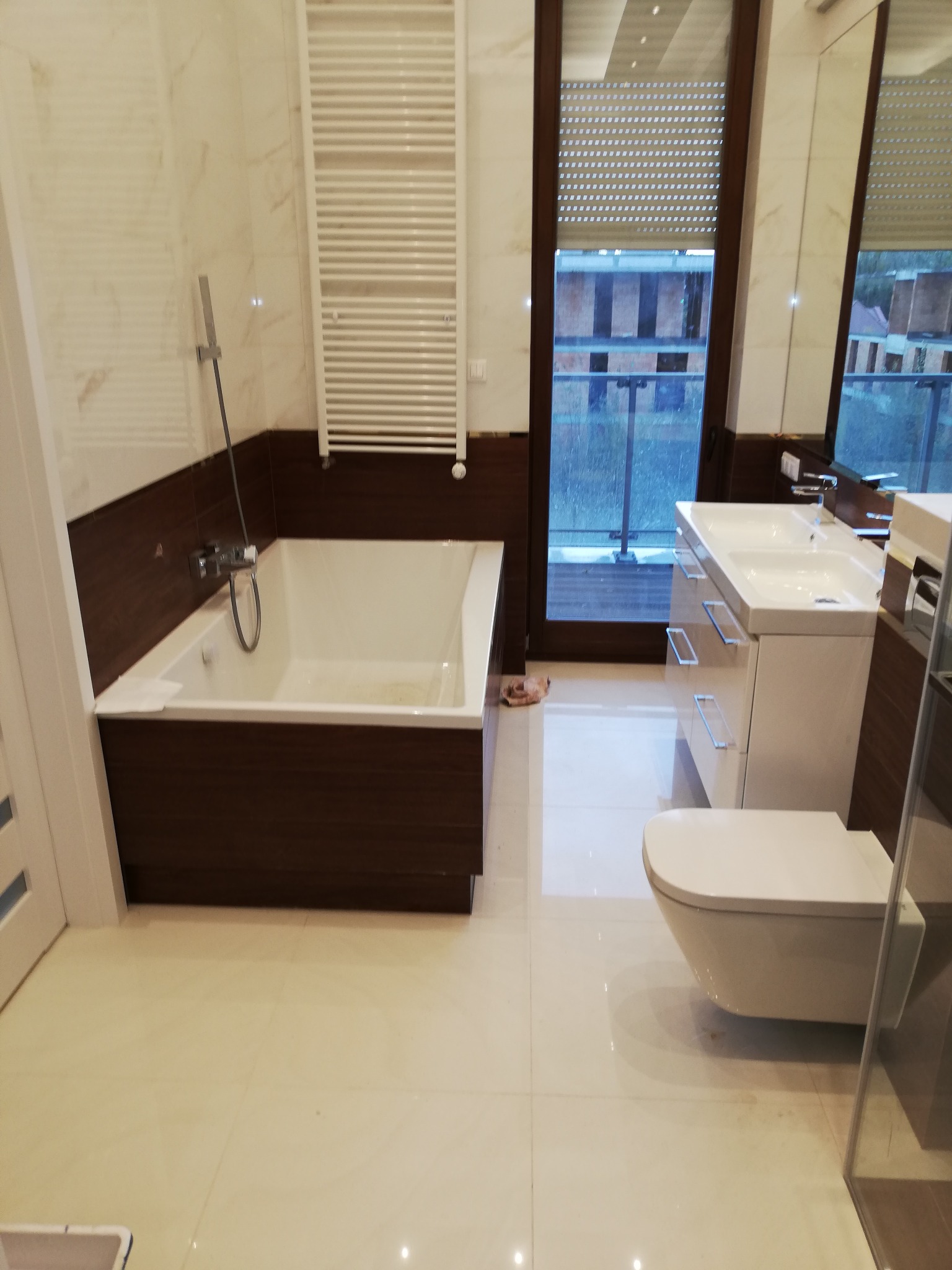 Nowoczesna łazienka z wanną w zabudowie z ciemnego drewna, umywalką z szafką, wiszącą toaletą i grzejnikiem drabinkowym na ścianie z marmurowym wzorem.