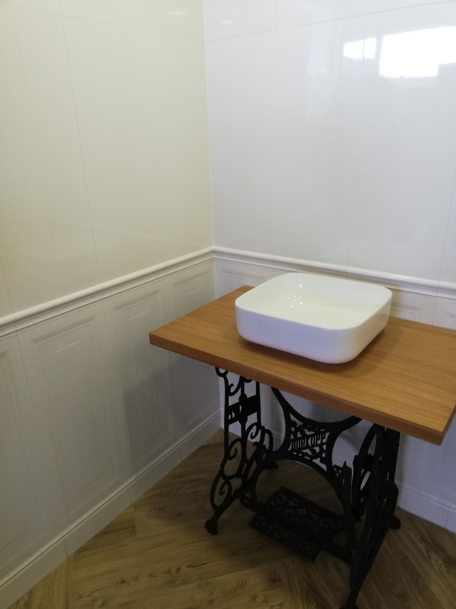 Biała, prostokątna umywalka nablatowa na drewnianym blacie, wsparta na żeliwnej, ozdobnej podstawie starej maszyny do szycia, na tle białych paneli ściennych z dekoracyjnymi listwami.