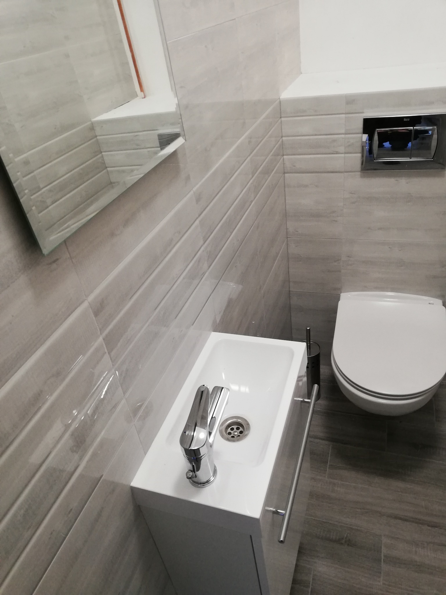 Nowoczesna łazienka w odcieniach szarości z podwieszaną toaletą, umywalką z szafką i dużym lustrem na ścianie z płytek imitujących drewno.