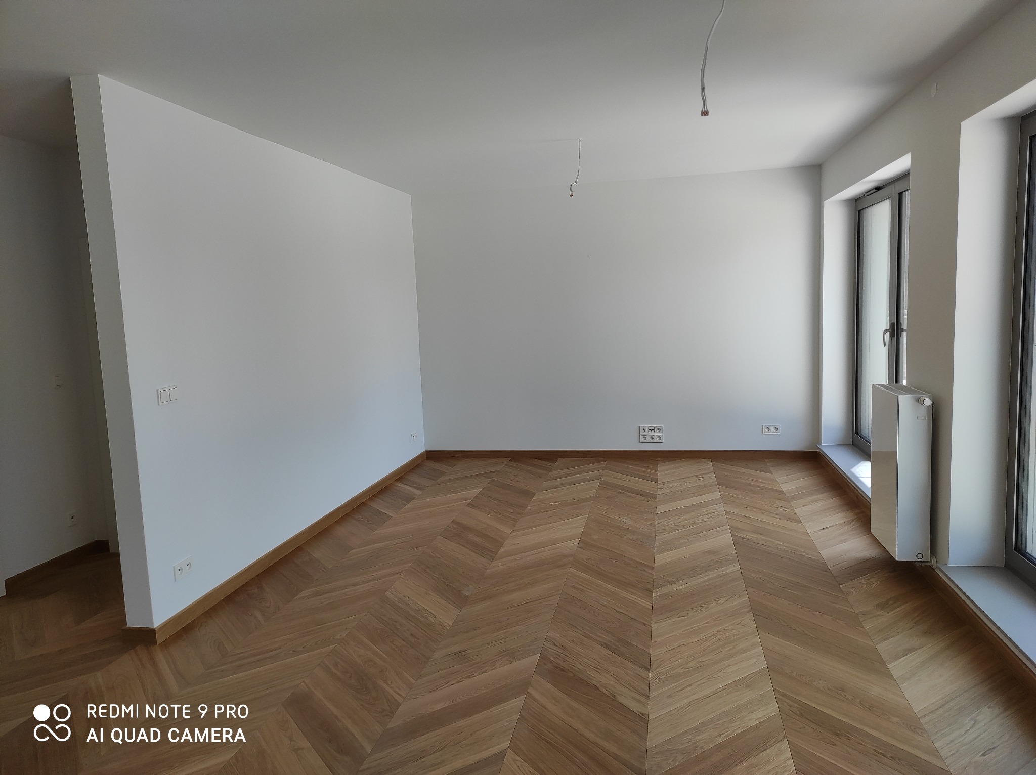 Pusty pokój z parkietem ułożonym w jodełkę, świeżo pomalowane białe ściany, widoczne gniazdka elektryczne i wystające przewody z sufitu, okno z grzejnikiem.