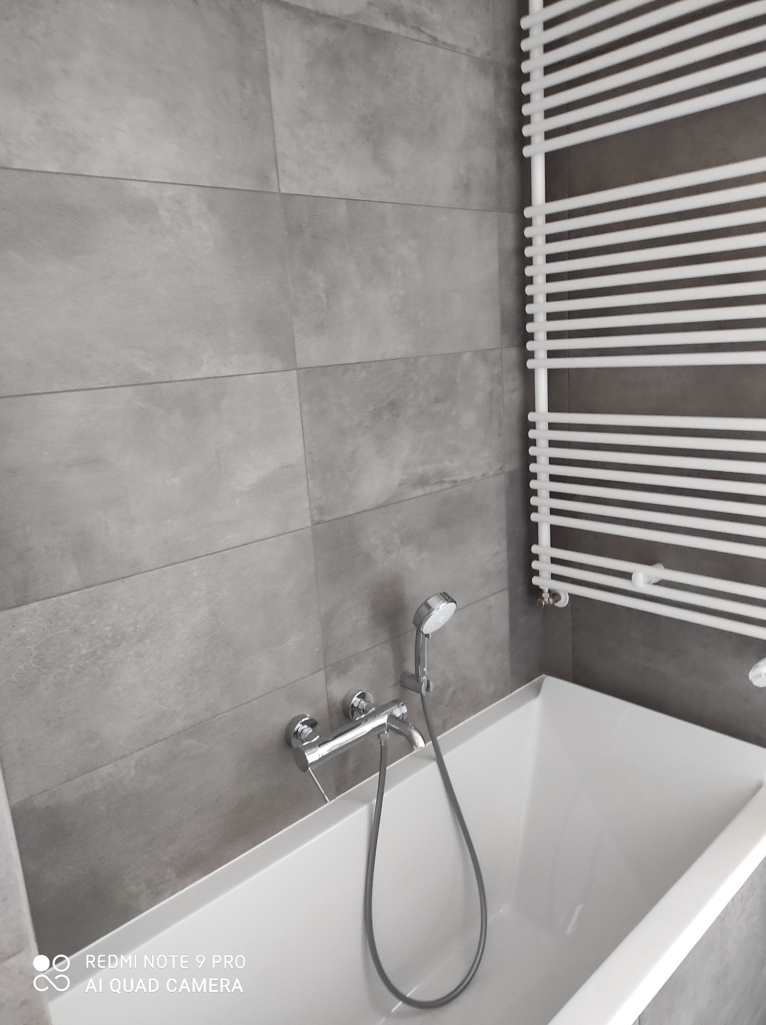 Nowoczesna łazienka z wanną, szarymi płytkami imitującymi beton i białym grzejnikiem drabinkowym.