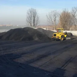Żółta ładowarka kołowa przy hałdach węgla na placu składowym, widoczne ślady opon na ziemi.
