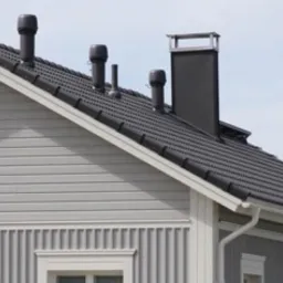 Szary dom jednorodzinny z ciemną dachówką i kilkoma kominami wentylacyjnymi o różnej wysokości.