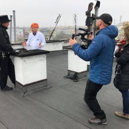 ZAKŁAD KOMINIARSKI KOMINIARCZYK - Ekipa filmowa kręci ujęcie na dachu budynku z kominiarzem w tradycyjnym stroju i mężczyzną w białym kitlu, prawdopodobnie podczas wywiadu lub scenki.