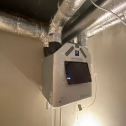 Centrala wentylacyjna z odzyskiem ciepła, model Aeris, z podłączonymi przewodami izolowanymi termicznie, częściowo owinięta folią ochronną, zamontowana przy suficie.