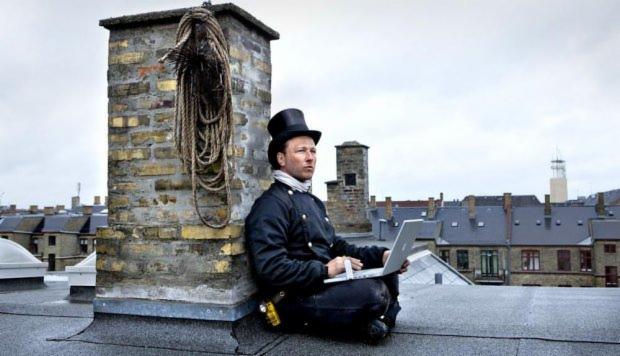 Osoba w tradycyjnym stroju, siedząca na dachu obok komina z laptopem, na tle miejskiego krajobrazu