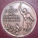 Srebrny medal 'Za Zasługi dla Rzemiosła Polskiego' z wizerunkiem rzemieślnika trzymającego miecz, na ciemnoczerwonym tle.