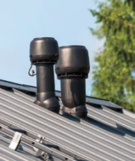 Dwa czarne kominki wentylacyjne na szarym, metalowym dachu, z widocznym fragmentem drabiny dekarskiej w lewym dolnym rogu, na tle błękitnego nieba i zielonych drzew.