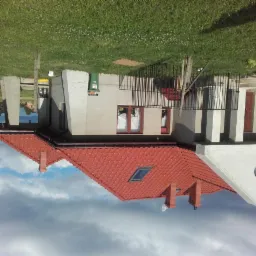 Dwupiętrowy dom z czerwonym dachem, widoczny z zewnątrz, z elementami nowoczesnej architektury, takimi jak pionowe, białe kolumny przy wejściu i okrągłe okno na szczycie ściany; na pierwszym planie...