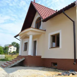 Elewacja domu jednorodzinnego w trakcie wykończenia, z widoczną ocieploną ścianą w kolorze beżowym, ceglaną podmurówką i schodami wejściowymi z płytek, na pierwszym planie piasek i siatka budowlana.