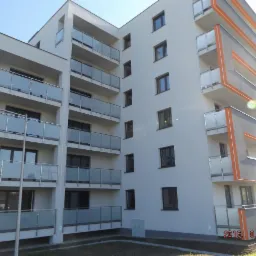 Nowoczesny budynek wielorodzinny z białymi elewacjami i balkonami z przezroczystymi balustradami, z pomarańczowymi akcentami na krawędziach balkonów i szarymi elementami dekoracyjnymi.