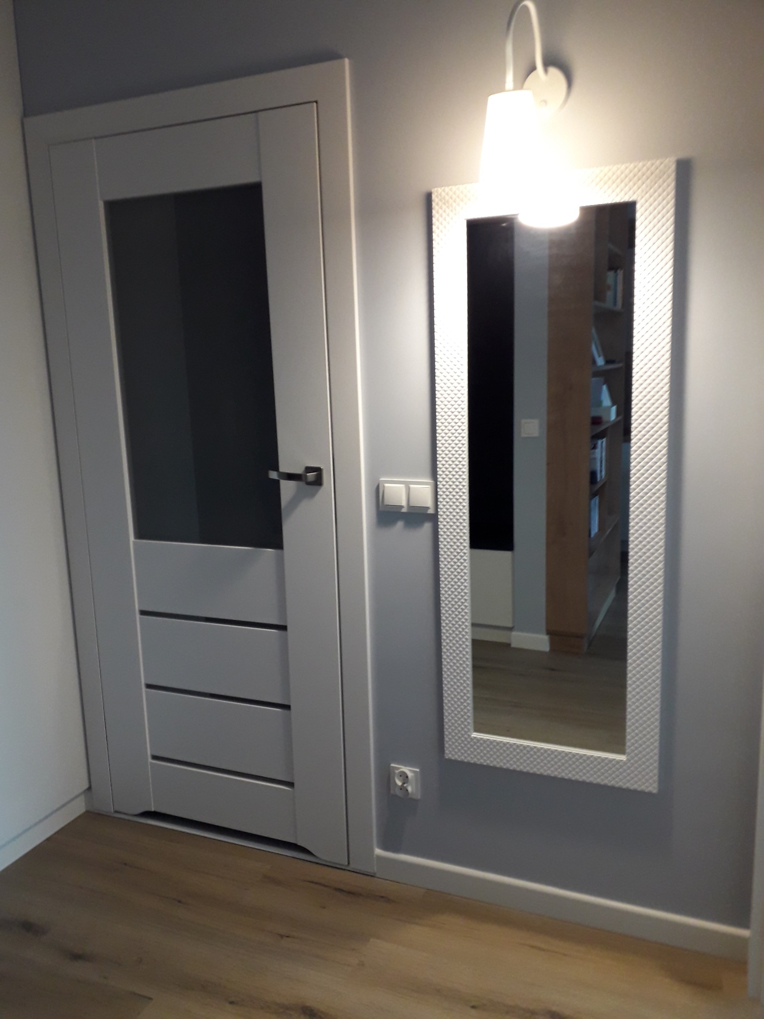 Szare drzwi wewnętrzne z przeszkleniem i poziomymi frezami, obok lustro w ozdobnej ramie oświetlone kinkietem, widok na wnętrze z regałem na książki.