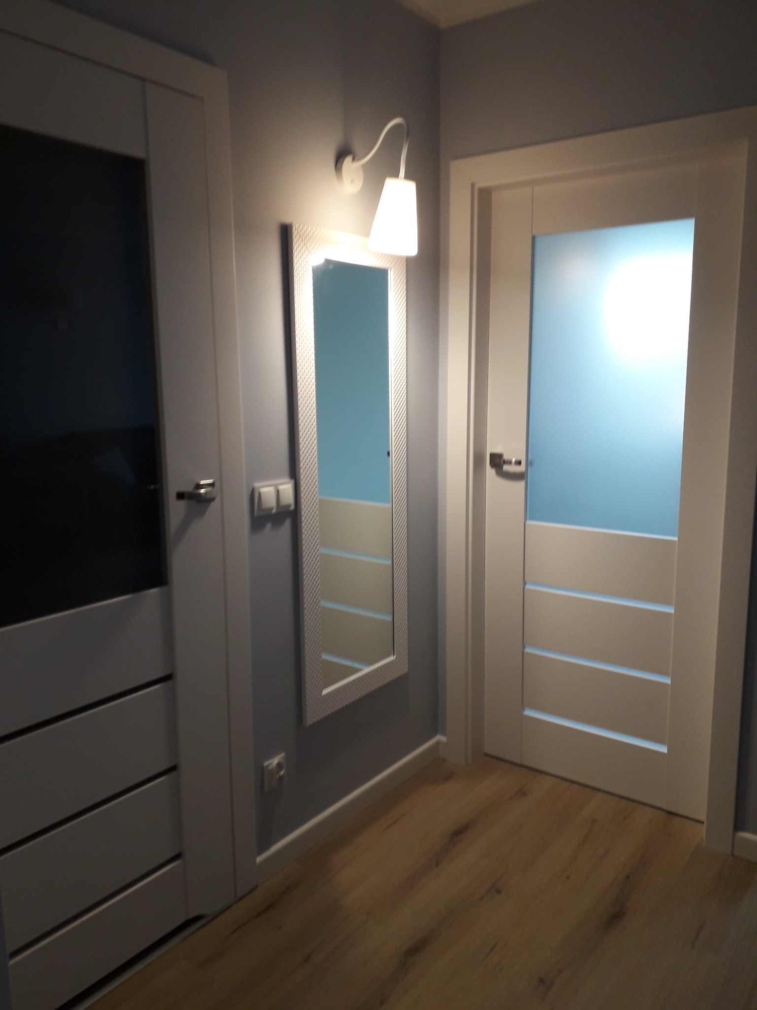 Wnętrze przedpokoju z dwoma białymi drzwiami z przeszkleniami, lustrem w ozdobnej ramie i drewnianą podłogą.