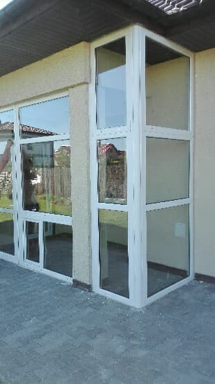 Nowoczesna zabudowa tarasu z białymi ramami okiennymi, widoczne refleksy w szybach i szara kostka brukowa na podłodze.