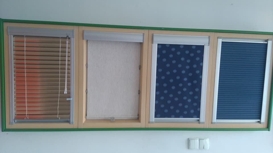Ekspozycja różnych typów rolet okiennych: drewniane żaluzje, kremowa roleta materiałowa, granatowa roleta w kropki i plisowana roleta w odcieniach niebieskiego, zamontowane w ramie z zielonym...