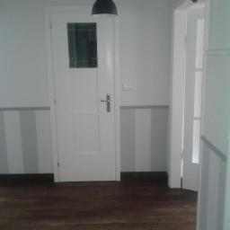 REM-BUDD Piotr Czachowski - Wnętrze po odświeżeniu: białe ściany z szarymi pasami, drewniana podłoga, białe drzwi z szybą i ościeżnica, lampa sufitowa. Minimalistyczny design.