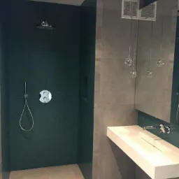 Nowoczesna łazienka z kabiną prysznicową wyłożoną ciemnozieloną mozaiką, wiszącą białą umywalką z chromowaną baterią i szklanymi lampami wiszącymi.