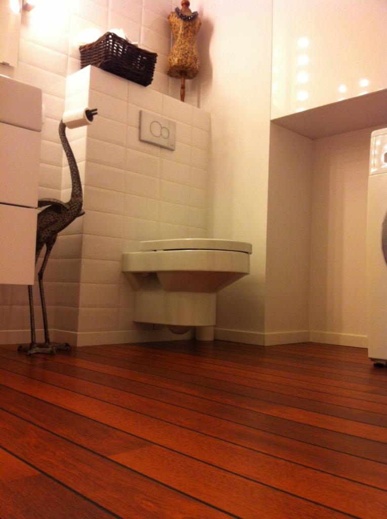 Nowoczesna łazienka z wiszącą toaletą, drewnianą podłogą, białymi płytkami i dekoracyjną figurką żurawia trzymającego papier toaletowy.