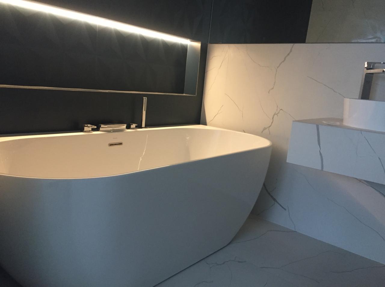 Nowoczesna, biała wanna wolnostojąca z chromowaną armaturą na tle ciemnej ściany z podświetleniem LED i marmurowej umywalki w wykończonej łazience w Warszawie.