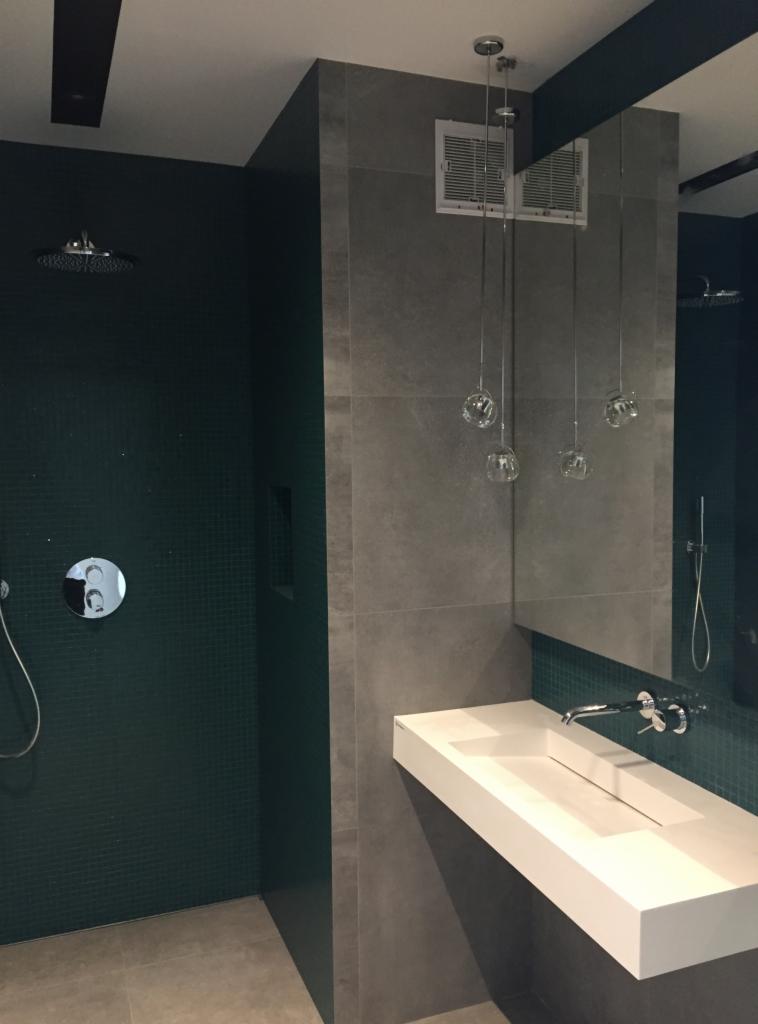 Nowoczesna łazienka z ciemnozieloną mozaiką pod prysznicem, szarą płytą wielkoformatową, wiszącą białą umywalką i designerskim oświetleniem w postaci szklanych kul na cienkich przewodach.