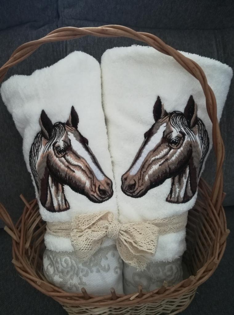 Dwa białe ręczniki z haftowanymi głowami koni, przewiązane koronkową wstążką i umieszczone w wiklinowym koszu.