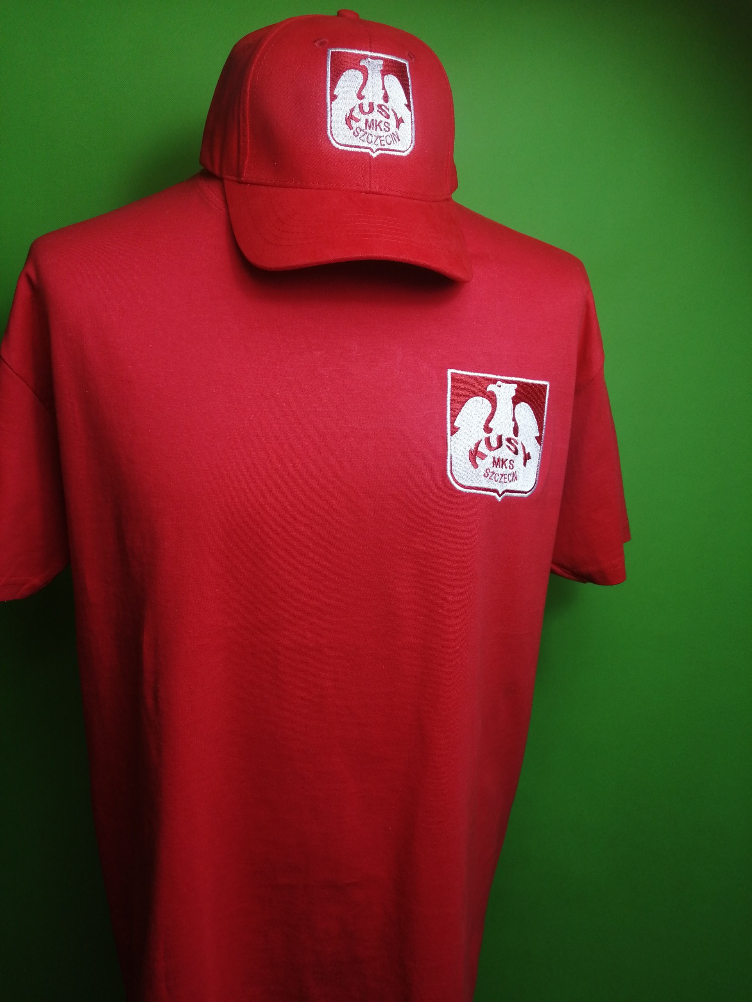 Czerwona koszulka i czapka z wyhaftowanym logo klubu sportowego MKS Szczecin na manekinie, na zielonym tle.