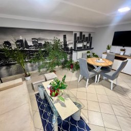 KSB Nieruchomości - Jasny salon z beżową sofą, szklanym stolikiem kawowym i okrągłym stołem. Na ścianie fototapeta z panoramą miasta. Wnętrze do kupna/wynajmu.