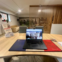 KSB Nieruchomości - Biuro nieruchomości: laptop z ofertą mieszkania na drewnianym biurku z podkładką w barwach Francji. W tle sofa, lamelki i grafika z panoramą miasta.