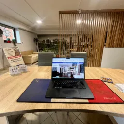 Biuro nieruchomości: laptop z ofertą mieszkania na drewnianym biurku z podkładką w barwach Francji. W tle sofa, lamelki i grafika z panoramą miasta.