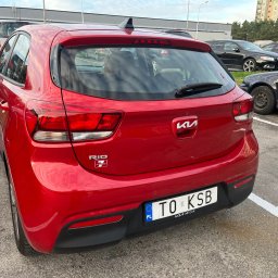 KSB Nieruchomości - Czerwone Kia Rio z tyłu, zaparkowane na parkingu, widoczny numer rejestracyjny i logo producenta. W tle inne samochody i budynki.