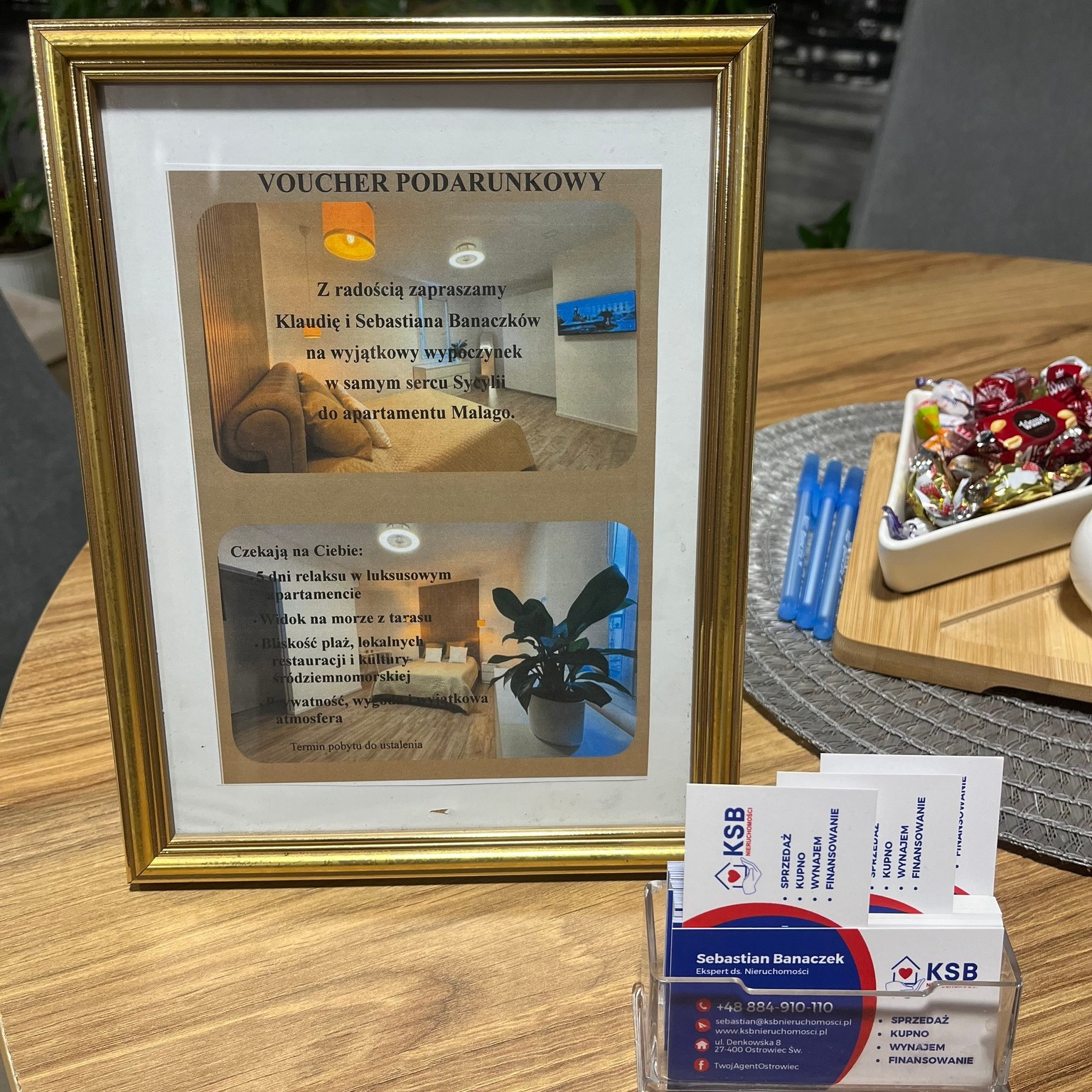 Voucher podarunkowy z ofertą apartamentu na Sycylii w ramce, wizytówki KSB Nieruchomości, słodycze i długopisy na drewnianym stole. Oferta wakacyjna.