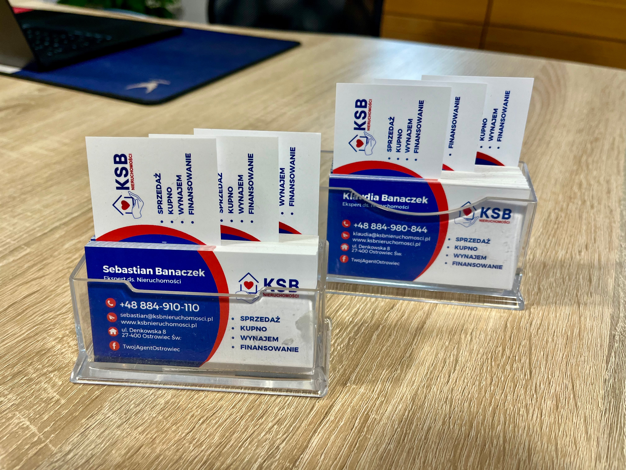Dwa przezroczyste stojaki z wizytówkami KSB Nieruchomości na drewnianym biurku, z ofertami sprzedaży, kupna, wynajmu i finansowania nieruchomości.