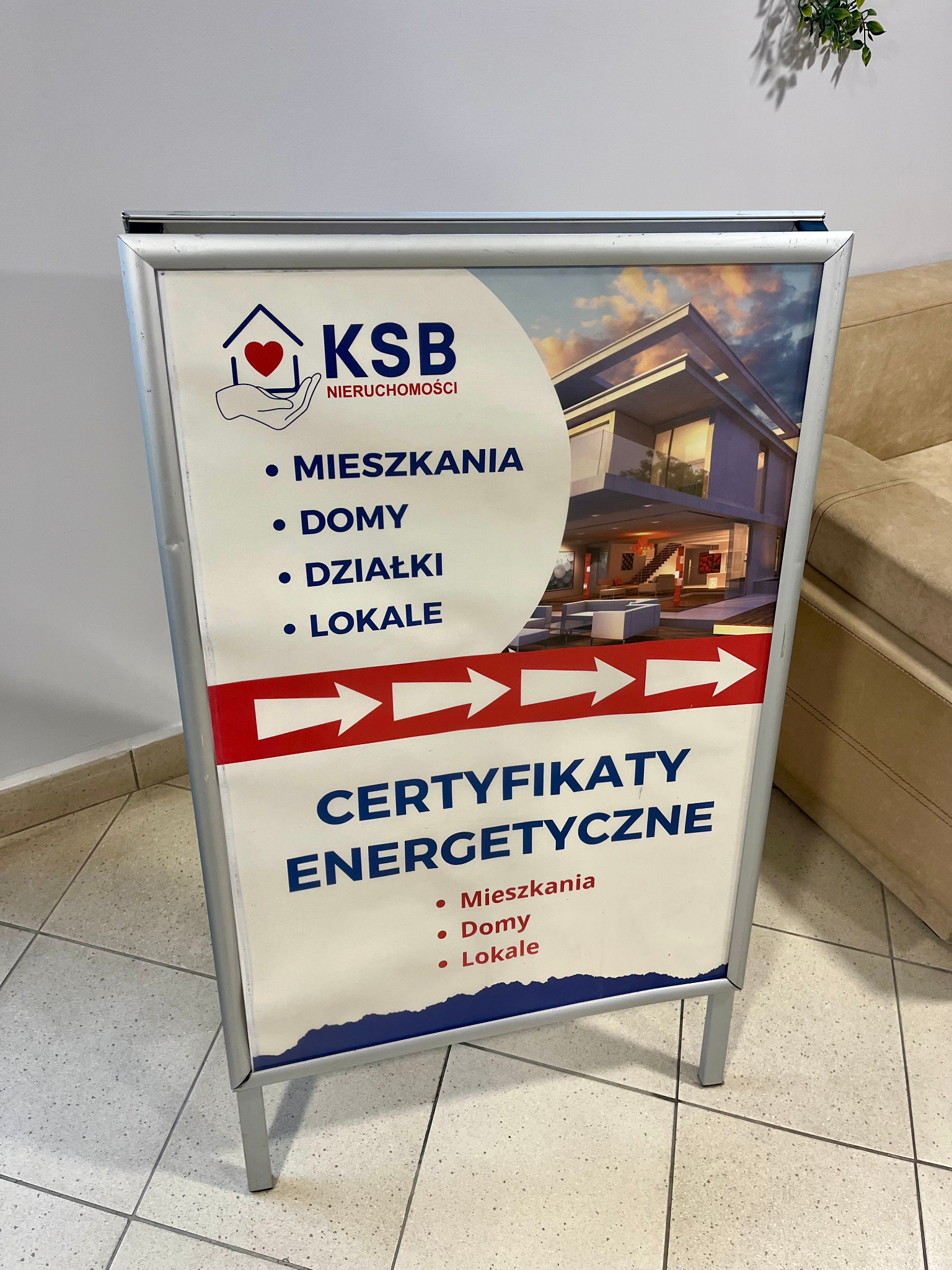 Stojak reklamowy KSB Nieruchomości z ofertą mieszkań, domów, działek i lokali oraz certyfikatów energetycznych. W tle nowoczesny dom.