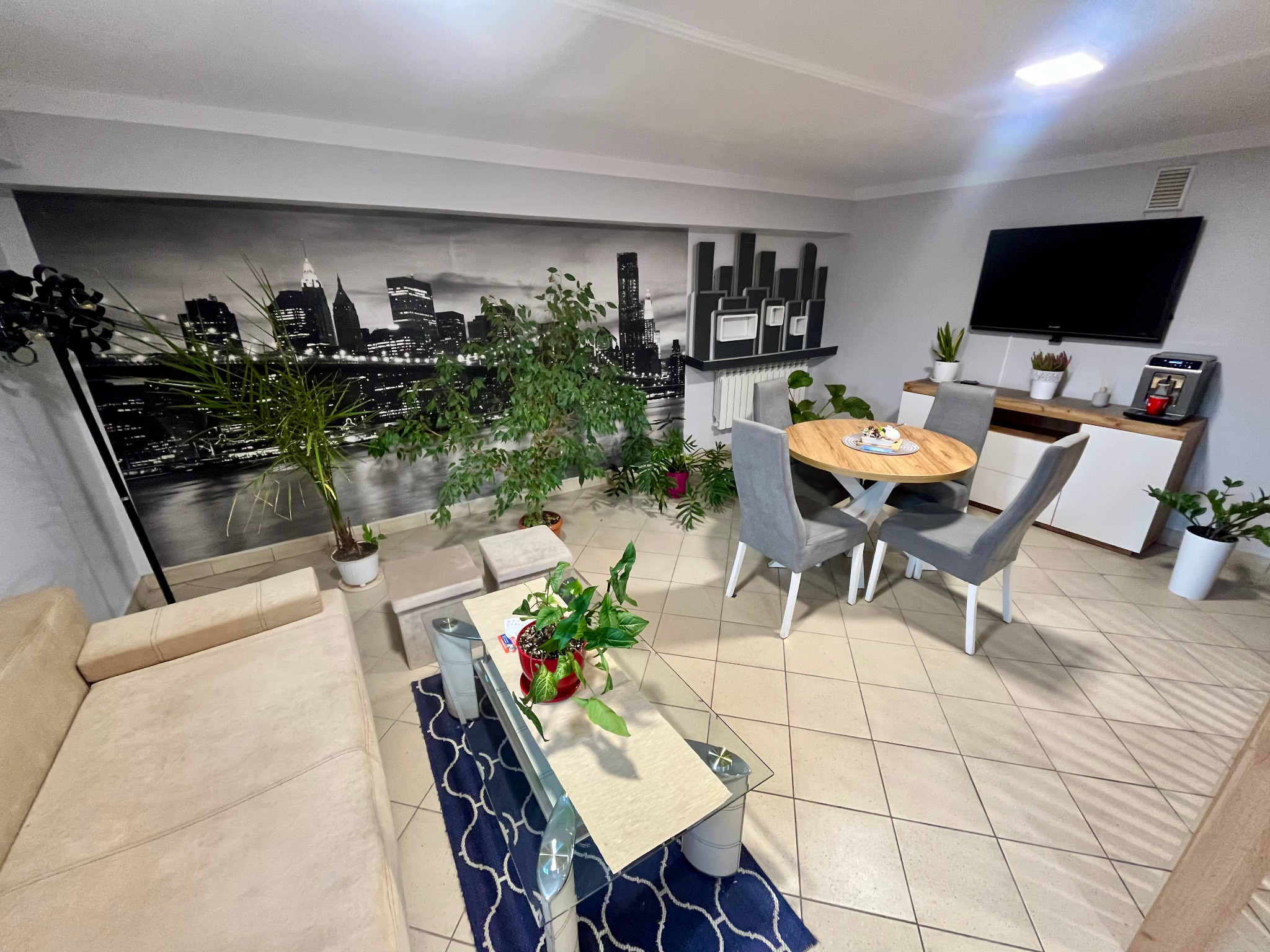 Jasny salon z beżową sofą, szklanym stolikiem kawowym i okrągłym stołem. Na ścianie fototapeta z panoramą miasta. Wnętrze do kupna/wynajmu.