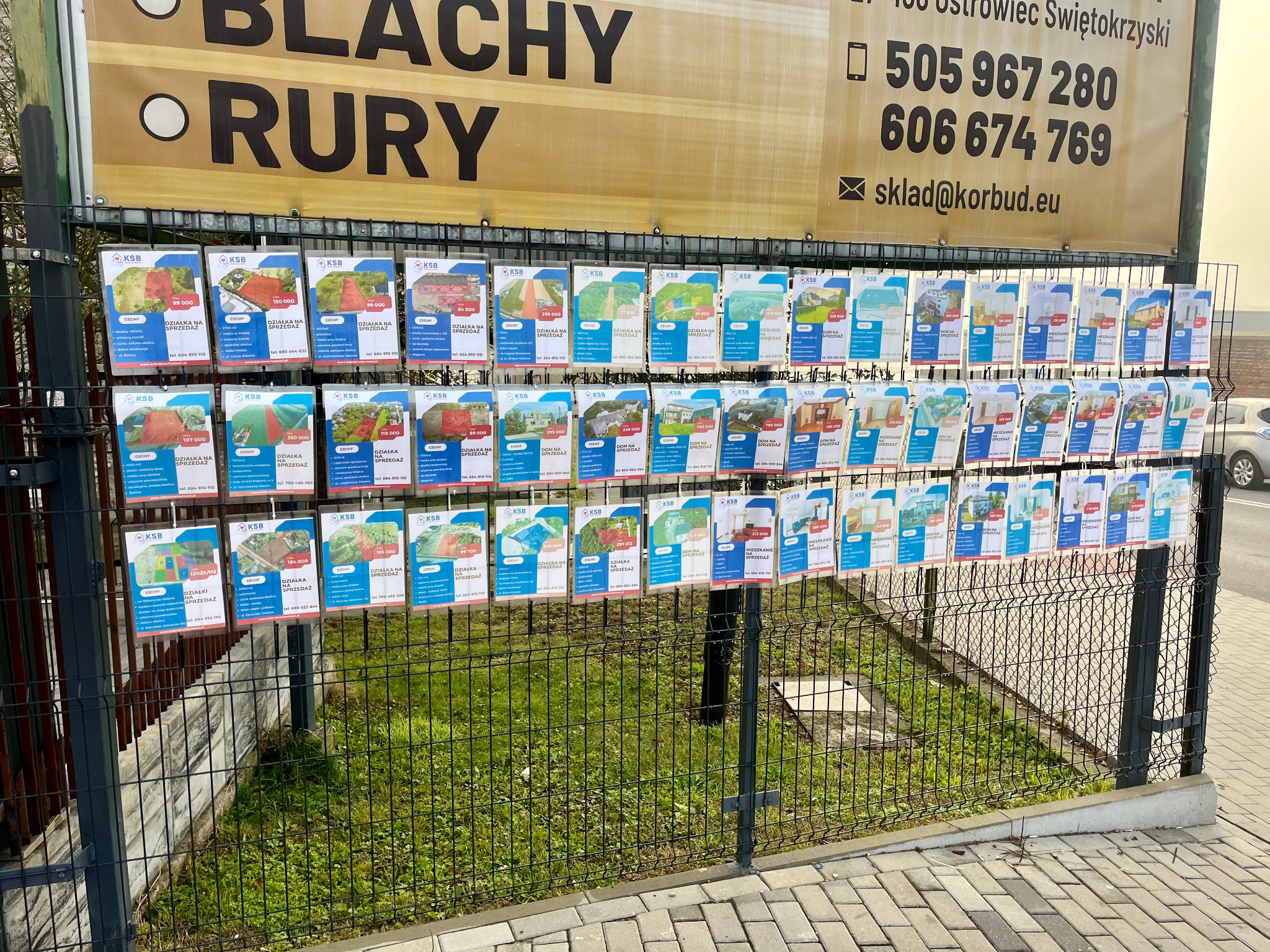 Ogrodzenie z ogłoszeniami 'Działka na sprzedaż', 'Dom na sprzedaż' i 'Mieszkanie na sprzedaż'. Reklama firmy KSB. Tablica informacyjna z numerami telefonów.