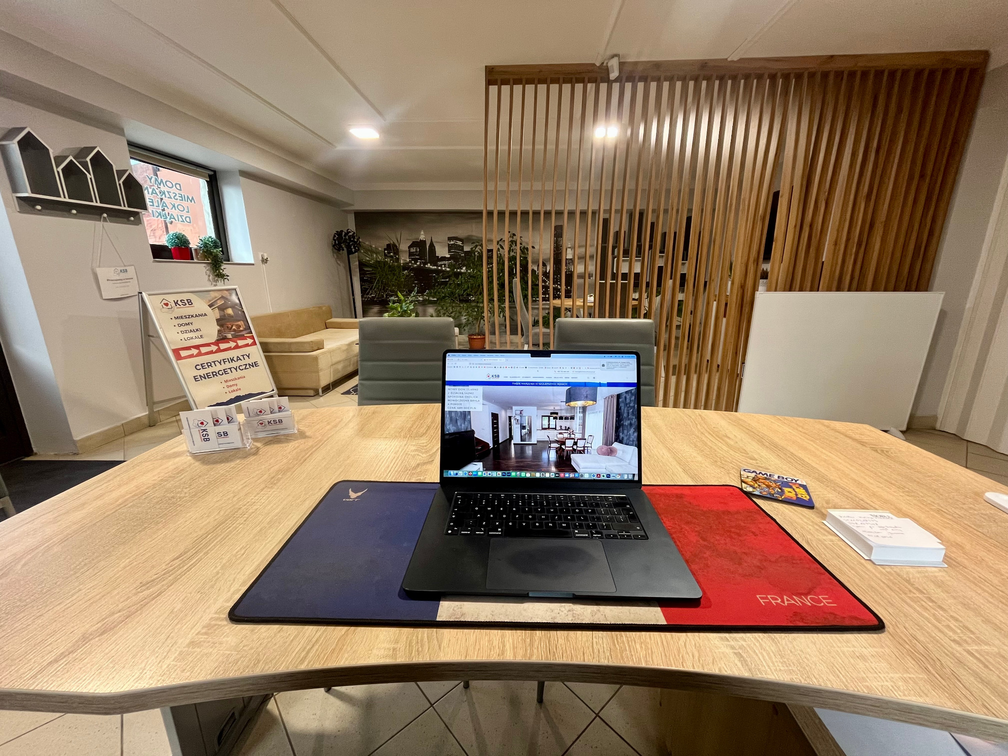 Biuro nieruchomości: laptop z ofertą mieszkania na drewnianym biurku z podkładką w barwach Francji. W tle sofa, lamelki i grafika z panoramą miasta.