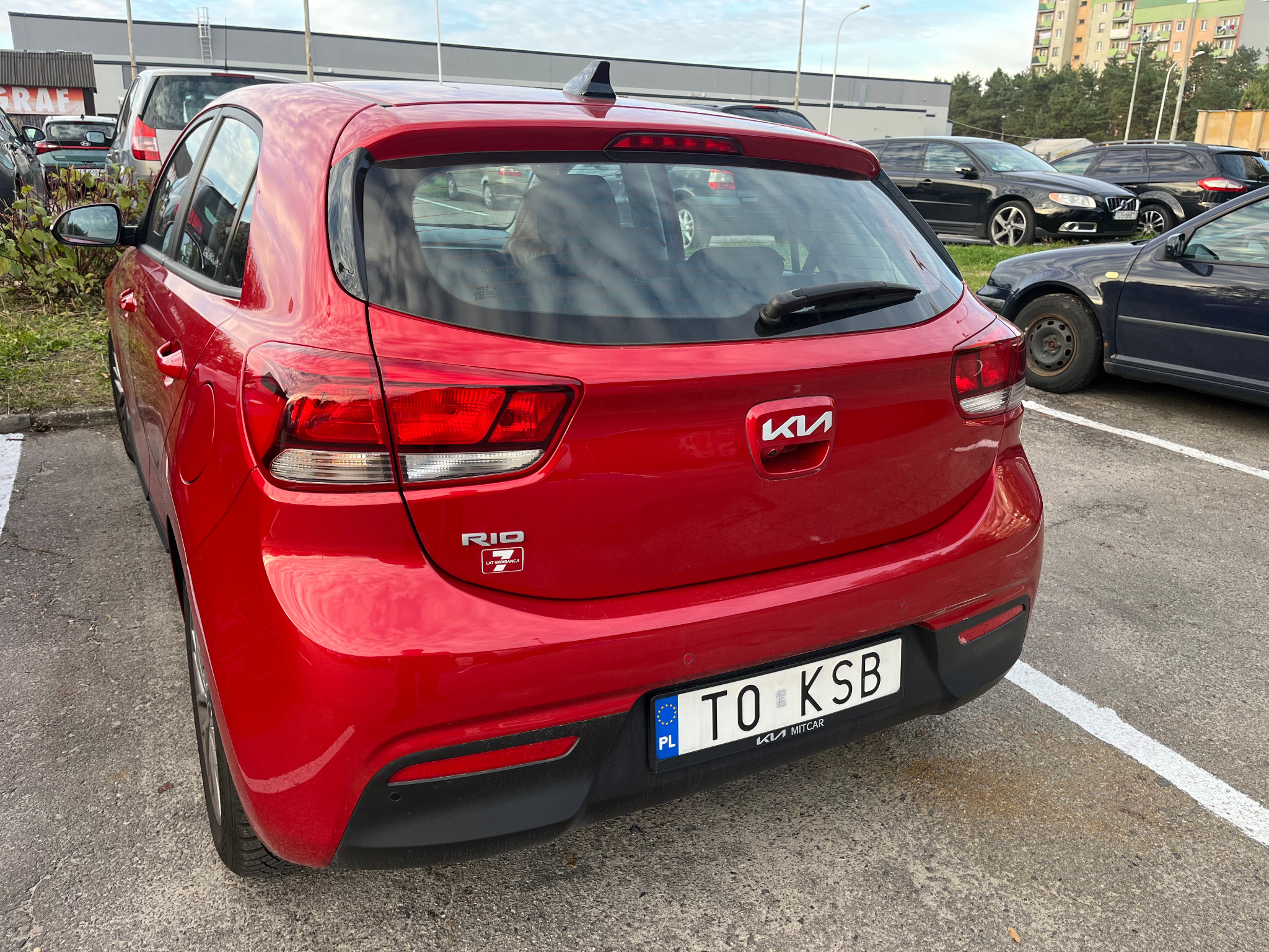 Czerwone Kia Rio z tyłu, zaparkowane na parkingu, widoczny numer rejestracyjny i logo producenta. W tle inne samochody i budynki.