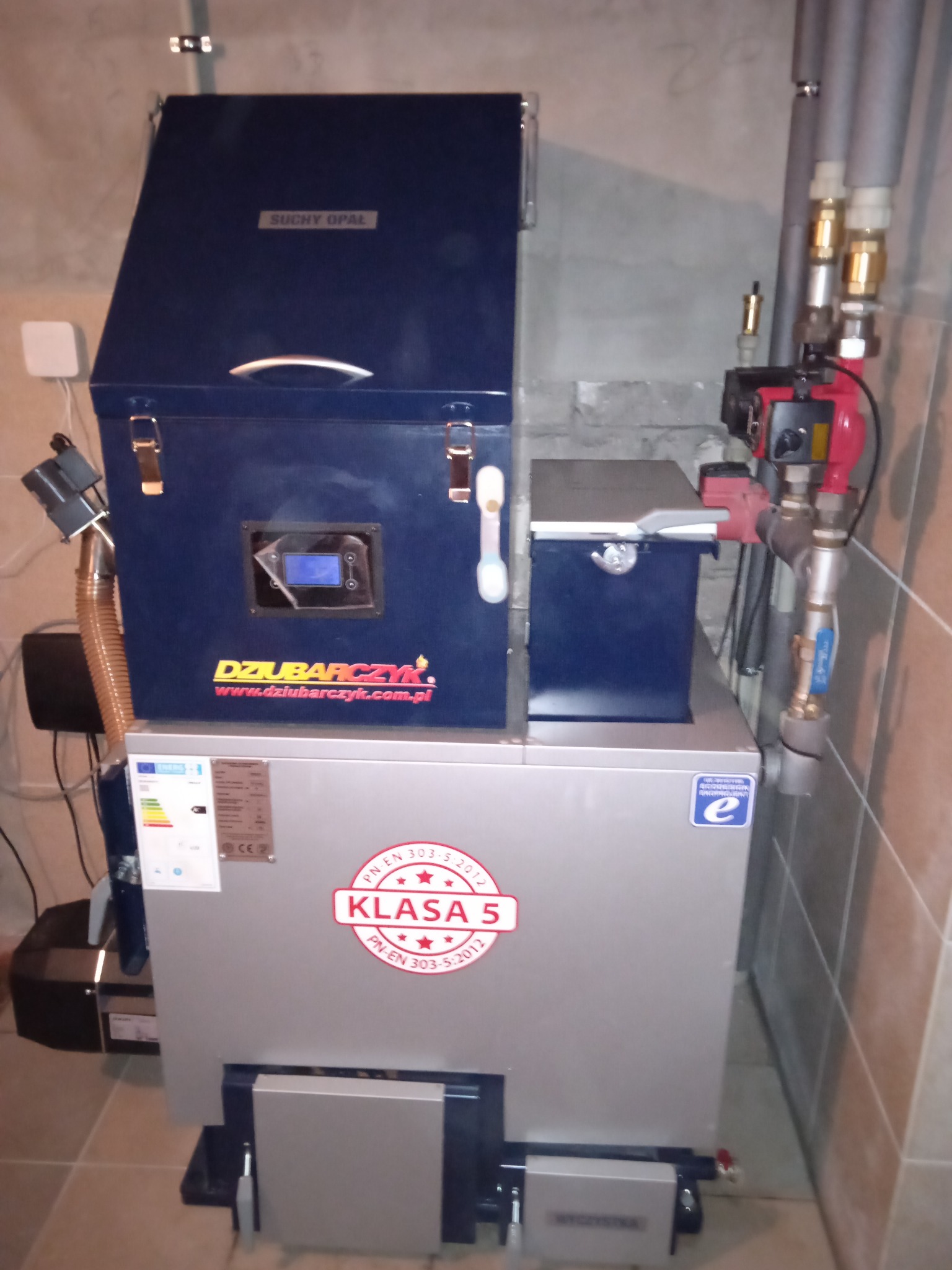 Nowoczesny kocioł na paliwo stałe marki Dziubarczyk, z widocznym oznaczeniem klasy 5, zainstalowany w kotłowni z wykończeniem z płytek.