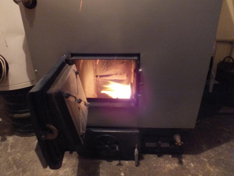 Otwarty piec na paliwo stałe z palącym się wewnątrz ogniem. Widoczna metalowa obudowa i fragment instalacji. Ujęcie z bliska, lekko z dołu.