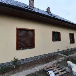 Elewacja domu jednorodzinnego w kolorze piaskowym, z brązowymi ramami okiennymi i roletą zewnętrzną, widoczne puszki z farbą i paleta na ziemi.