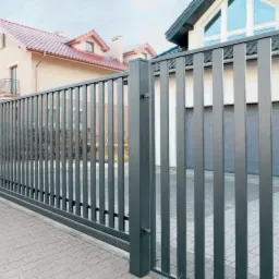 Nowoczesne, metalowe ogrodzenie panelowe w kolorze grafitowym, zamontowane przed domem w Rakoniewicach, z widoczną bramą wjazdową i kostką brukową.