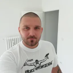 Mężczyzna w białej koszulce z logo firmy remontowej na tle świeżo pomalowanego, jasnego wnętrza. Widoczne drzwi i grzejnik.