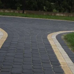 Ułożona, ciemnoszara kostka brukowa Nostalit z kolorowymi, kremowo-żółtymi pasami na obrzeżach, tworząca rozwidlenie ścieżki.