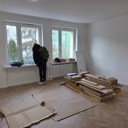 Usługi ogólnobudowlane Groździej Arkadiusz - Remont pokoju: parkiet w jodełkę, okna z widokiem na blok, kartony z panelami i osoba z plecakiem. Przygotowanie do montażu podłogi.