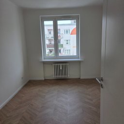 Usługi ogólnobudowlane Groździej Arkadiusz - Jasne wnętrze pokoju po remoncie: parkiet w jodełkę, białe ściany, okno z widokiem na budynek. Minimalistyczny grzejnik pod oknem. Otwarta biała futryna drzwi.