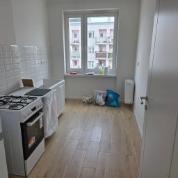 Usługi ogólnobudowlane Groździej Arkadiusz - Jasna kuchnia w bloku po remoncie: białe płytki, drewnopodobne panele, okno z widokiem na budynek. Nowa kuchenka gazowa i worki z materiałami budowlanymi.