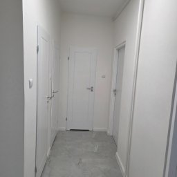 Usługi ogólnobudowlane Groździej Arkadiusz - Biały korytarz z trzema parami białych drzwi i szarą marmurową podłogą. Widoczne rury przy suficie. Minimalistyczny design.